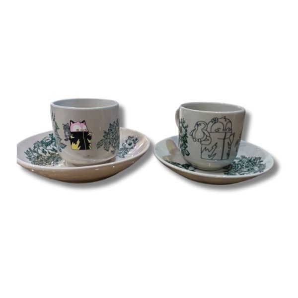 Two ceramic cups with unique designs on matching saucers: one features a pink cat with a black book, and the other displays a whimsical illustration of a bird and flowers.